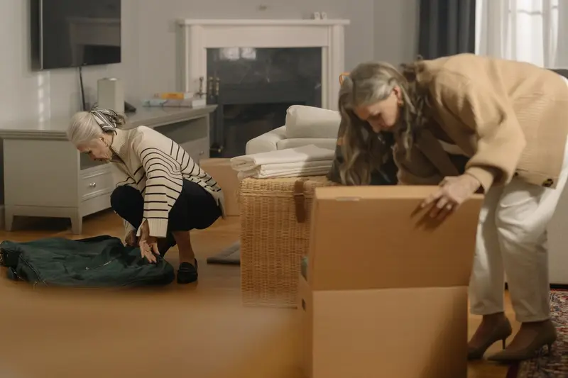 Two older women pack belongings into cardboard boxes in a living room