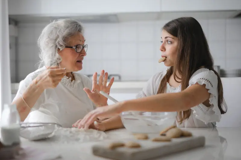 A grandmother and her granddaughter talk and laugh while baking cookies together in a bright kitchen