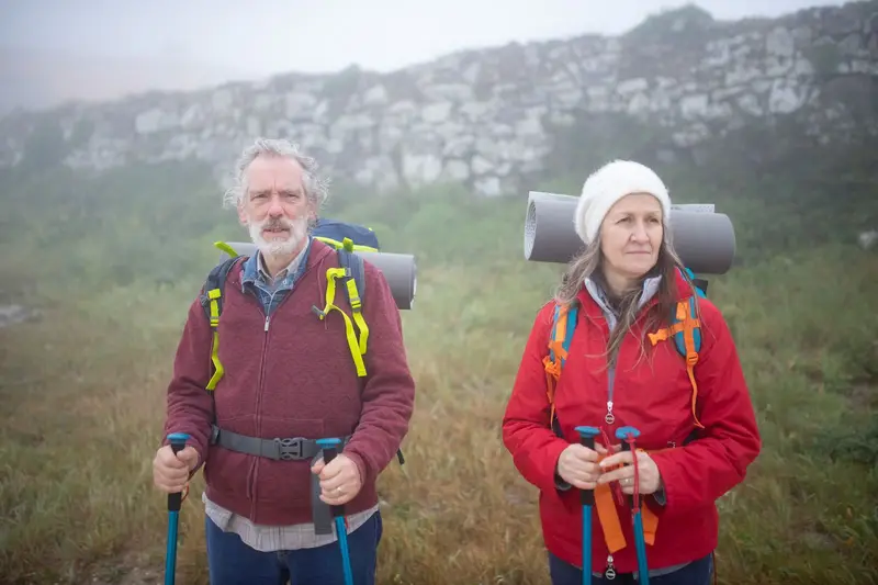 An older couple in rain jackets and backpacks smiles on a hiking trail